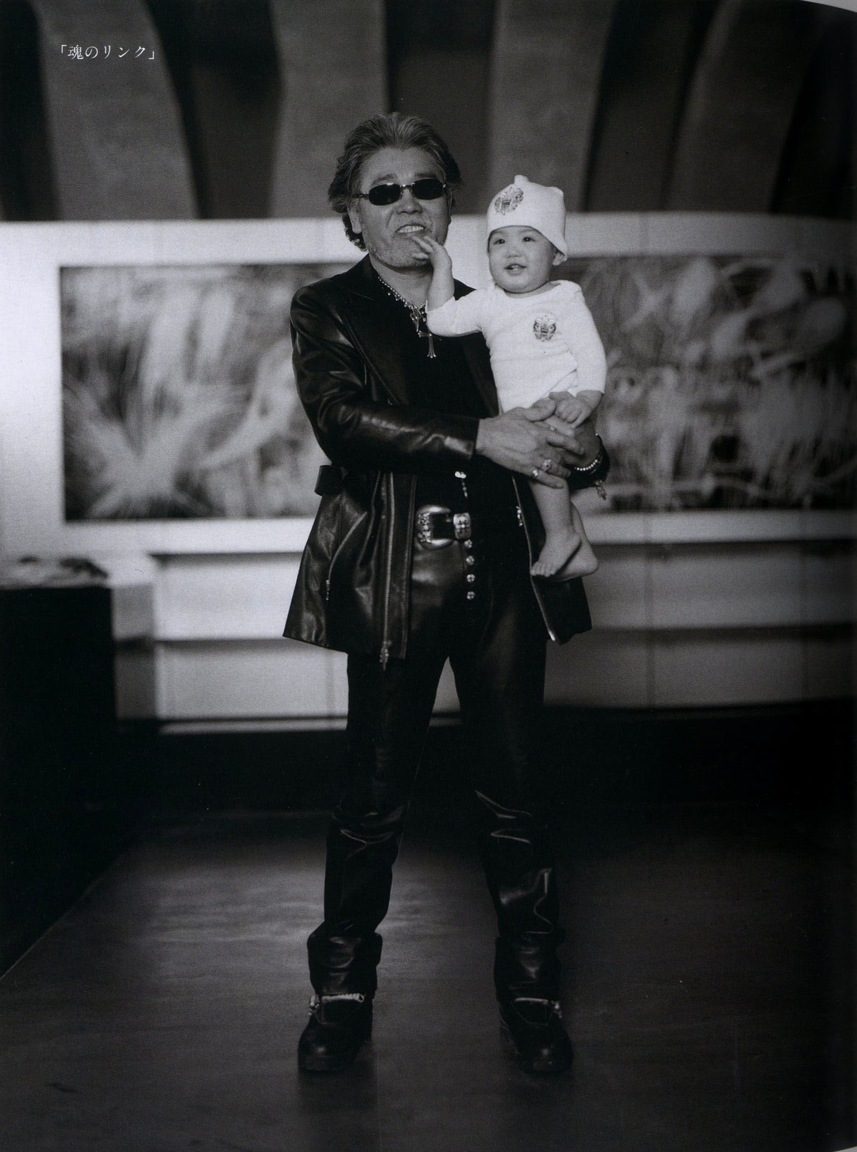 Black-and-white portrait of a man in a leather jacket holding a baby in front of a large artwork, Chrome Hearts Magazine Vol. 1.