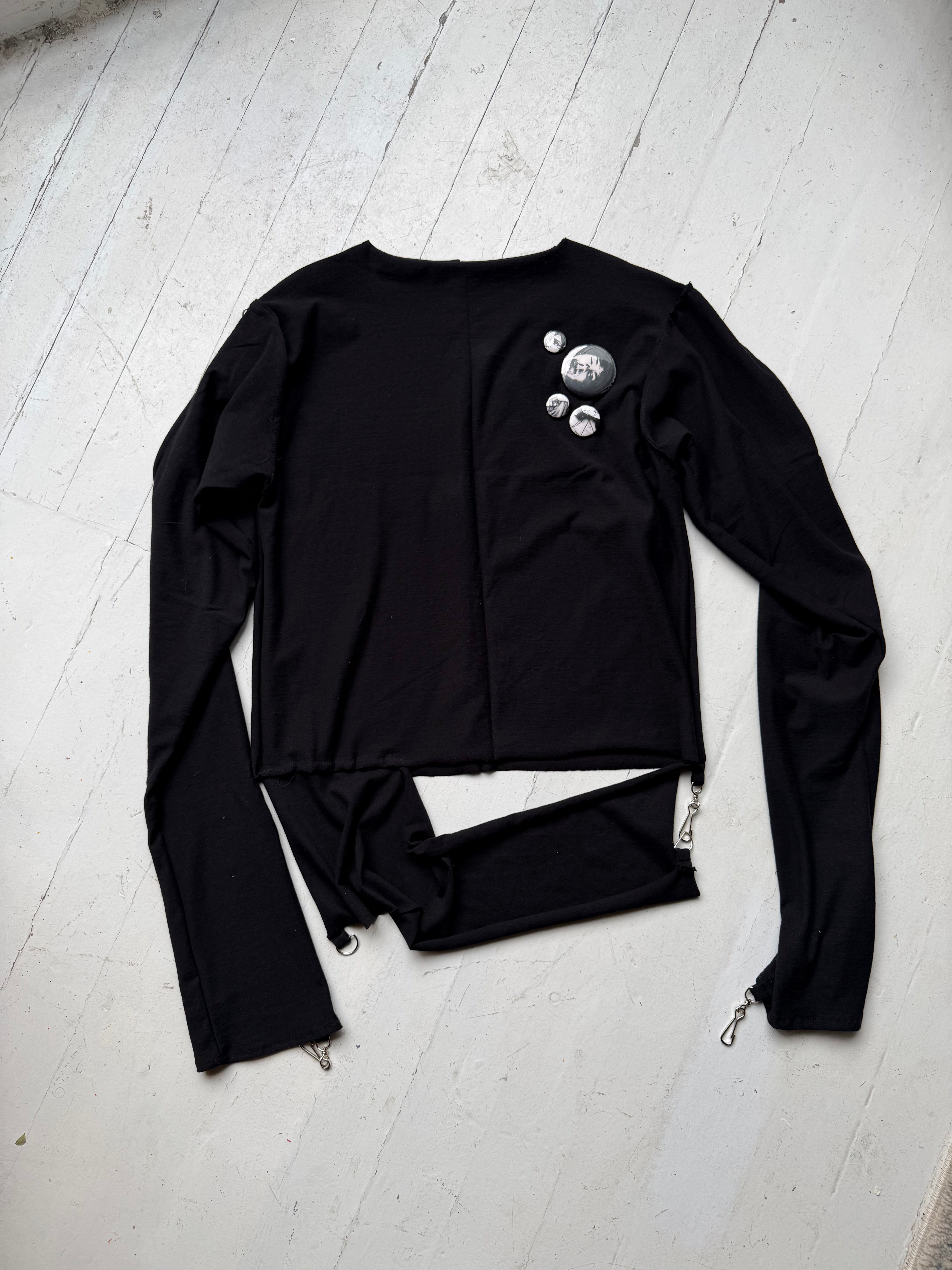 Black long-sleeve top by intermission with three pin-back buttons on the chest and a split, cut hem, laid flat.