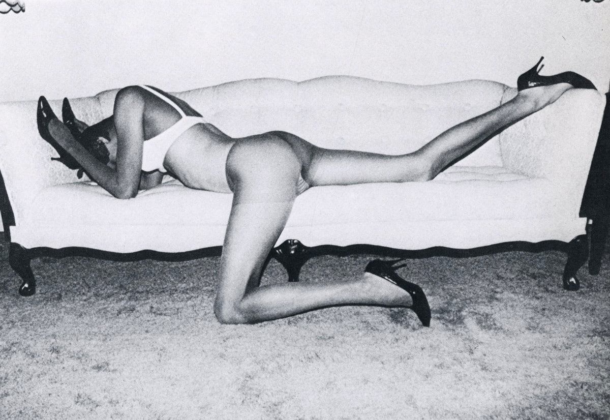 Black-and-white photograph from Submission: figure in high heels on all fours on a carpet before a white sofa, one leg extended.