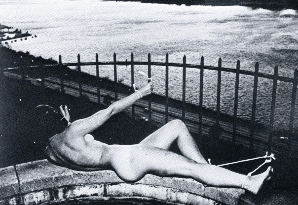 Jimmy DeSana photograph from Submission: nude figure lying on a stone ledge by an iron railing above the sea, a cord tied at the ankle.