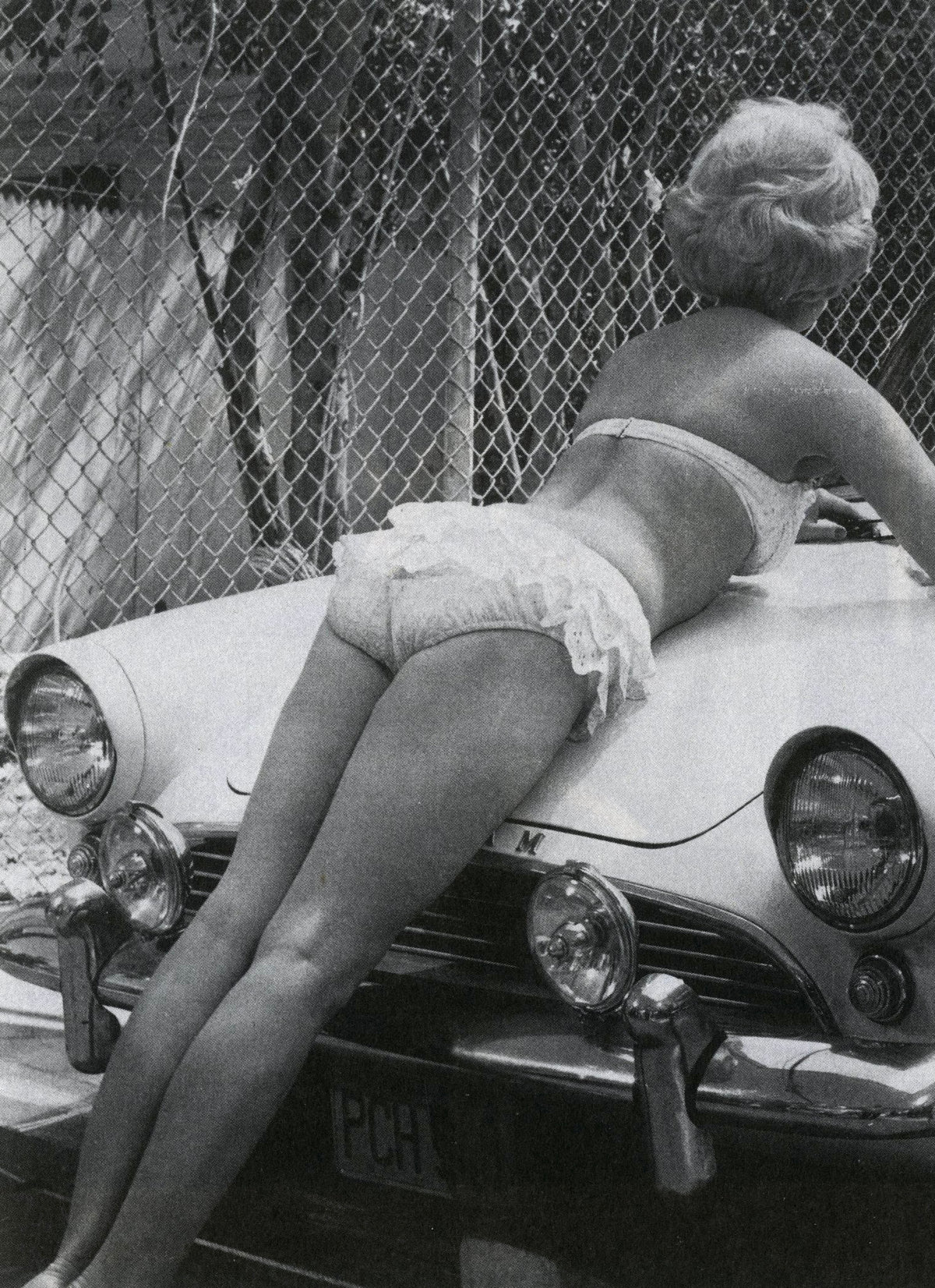 Woman in a bikini leaning over the hood of a vintage car by a chain-link fence, image from Fama & Fortune Heft 8.