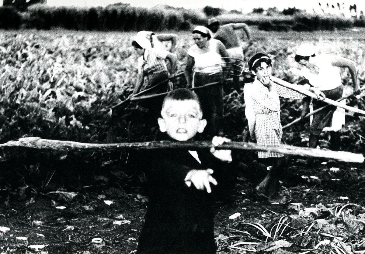 Photograph by Mario Giacomelli: a group of children in a field, one boy in the foreground holding a stick.